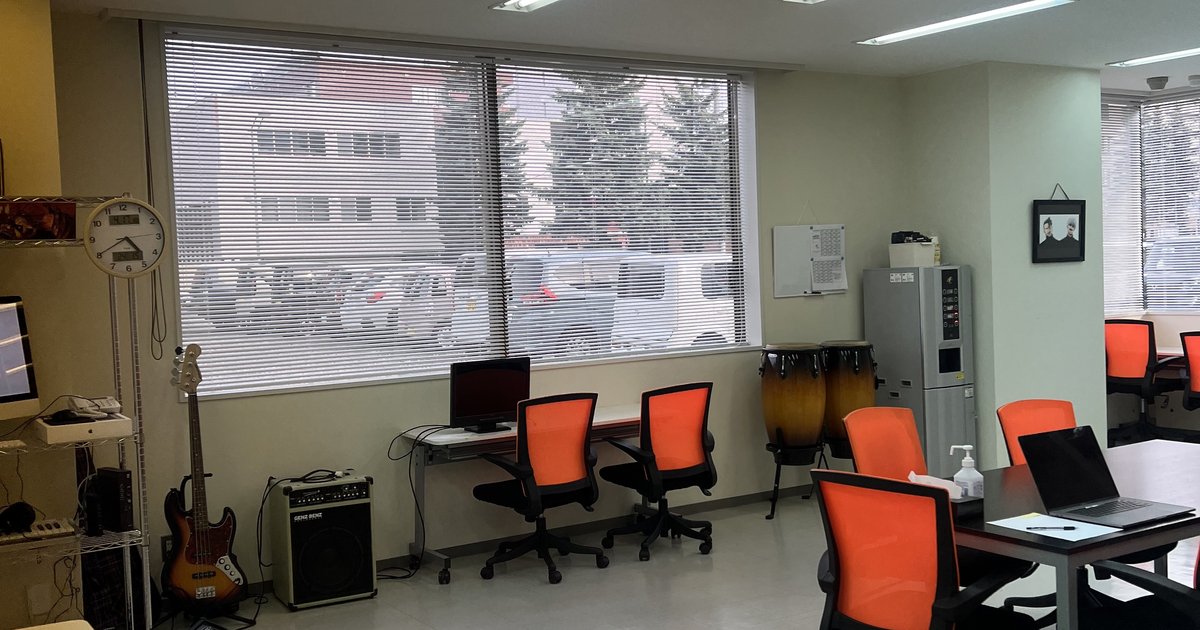 ALII旭川の明るい作業スペース。大きな窓から陽射しが入り、オレンジのチェア、ベースやコンガなどの楽器、ドリンクサーバーが見える